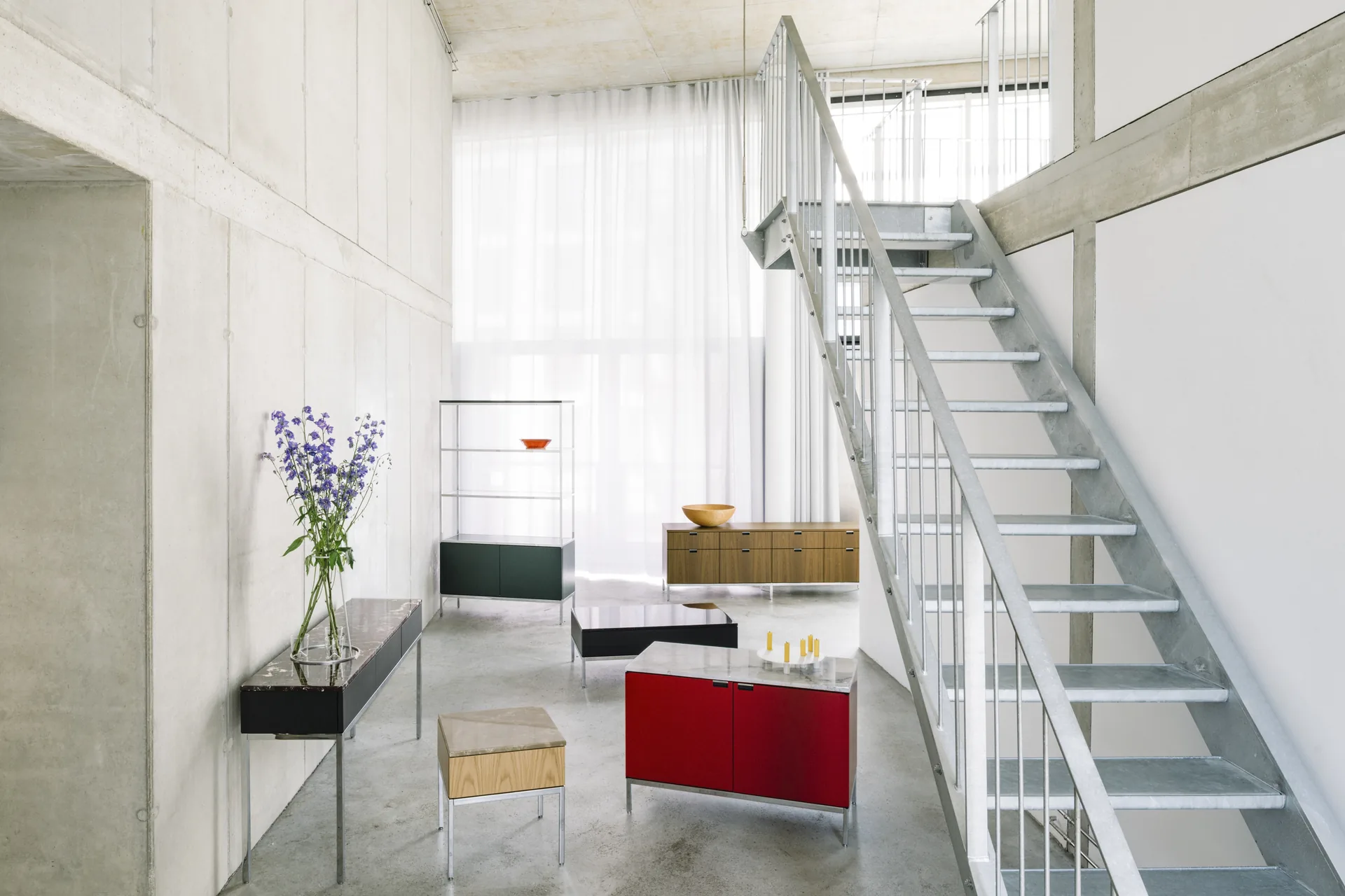 Knoll Chermayeff Storage: rotes Sideboard, schwarze Konsole und offenes Regal in einem Betonloft mit Stahltreppe